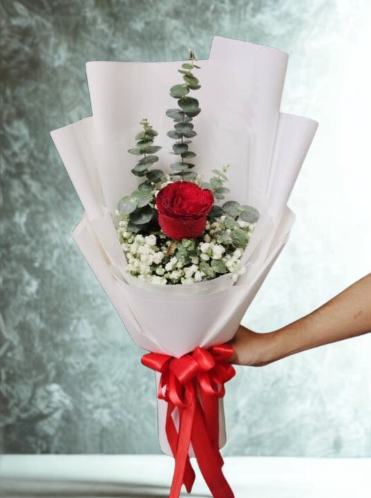 A minimalist single-rose bouquet with a bold romantic feel. One deep red rose stands at the center, softened by delicate white baby’s breath and fresh eucalyptus. Wrapped in clean white paper and finished with a bright red ribbon — simple, elegant, and perfect for a meaningful gesture.